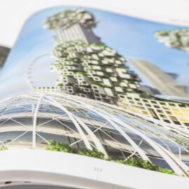 Das Bild zeigt ein geöffnetes Buch mit einer Darstellung futuristischer Architektur. Zu sehen sind hohe Gebäude mit ungewöhnlichen, geometrischen Formen und integrierten Grünflächen. Im Vordergrund befindet sich eine große Glaskuppelstruktur.