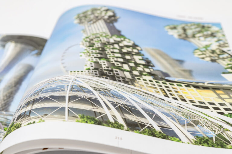 Das Bild zeigt ein geöffnetes Buch mit einer Darstellung futuristischer Architektur. Zu sehen sind hohe Gebäude mit ungewöhnlichen, geometrischen Formen und integrierten Grünflächen. Im Vordergrund befindet sich eine große Glaskuppelstruktur.