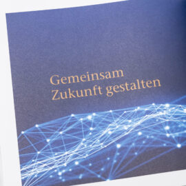 Das Bild zeigt ein geöffnetes Buch mit dem zentralen Text „Gemeinsam Zukunft gestalten“. Der Hintergrund ist dunkelblau und am unteren Rand verläuft ein netzartiges Muster aus hellblauen und weißen Linien und Punkten.