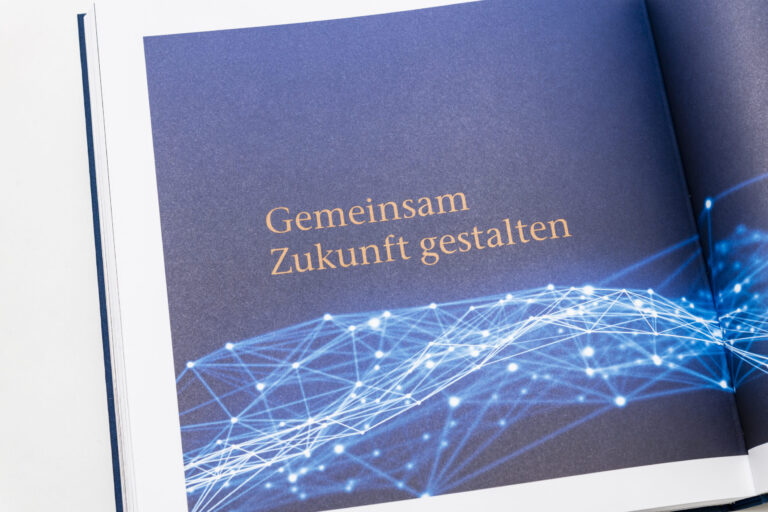 Das Bild zeigt ein geöffnetes Buch mit dem zentralen Text „Gemeinsam Zukunft gestalten“. Der Hintergrund ist dunkelblau und am unteren Rand verläuft ein netzartiges Muster aus hellblauen und weißen Linien und Punkten.