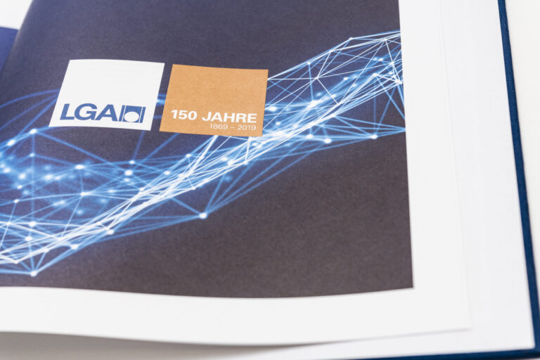 Das Bild zeigt eine Seite aus einem Buch oder Magazin mit dunklem Hintergrund und einem abstrakten Muster aus blauen Linien und Punkten. In der Mitte befinden sich zwei farbige Quadrate: ein weißes Quadrat mit der Aufschrift „LGA“ und geometrischem Logo sowie ein bronzenes Quadrat mit der Aufschrift „150 JAHRE 1869 – 2019“.