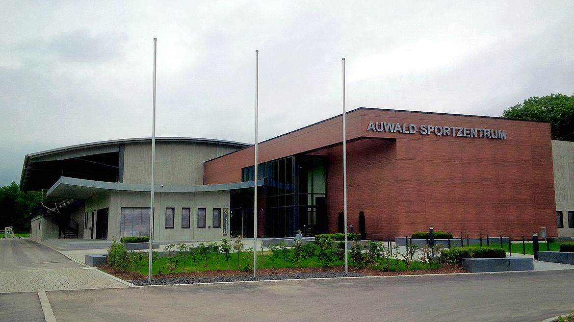 Modernes Sportzentrum mit der Aufschrift ‚AUWALD SPORTZENTRUM‘ an der Fassade, kombiniert aus Beton, Ziegel und großen Glasflächen; davor drei Fahnenmasten.