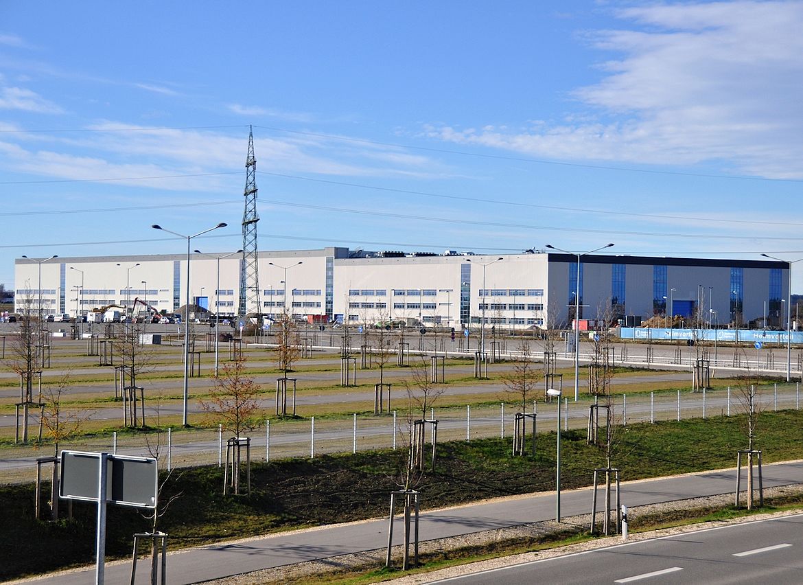Ein großes Industrie- oder Lagergebäude mit mehreren Laderampen und Parkflächen steht hinter einer Reihe junger Bäume und Straßenlaternen entlang einer parallel verlaufenden Straße; im Hintergrund ist ein hoher Strommast unter einem überwiegend klaren Himmel mit einigen Wolken zu sehen.