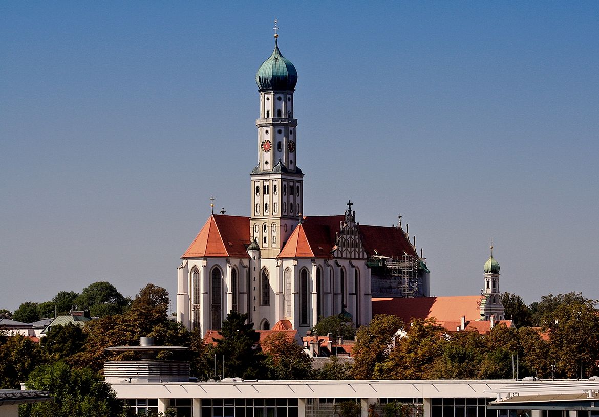 Historische Kirche mit hohem zentralen Turm und grünem Kuppeldach, umgeben von roten Ziegeldächern, Bäumen und weiteren Gebäuden.