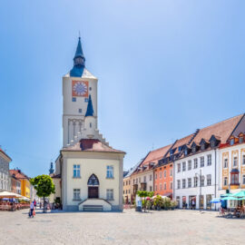 Belebter Stadtplatz in Deggendorf mit einem hohen Uhrturm und bunten Gebäuden, umgeben von Außengastronomie mit Sonnenschirmen und Spaziergängern bei sonnigem Wetter.