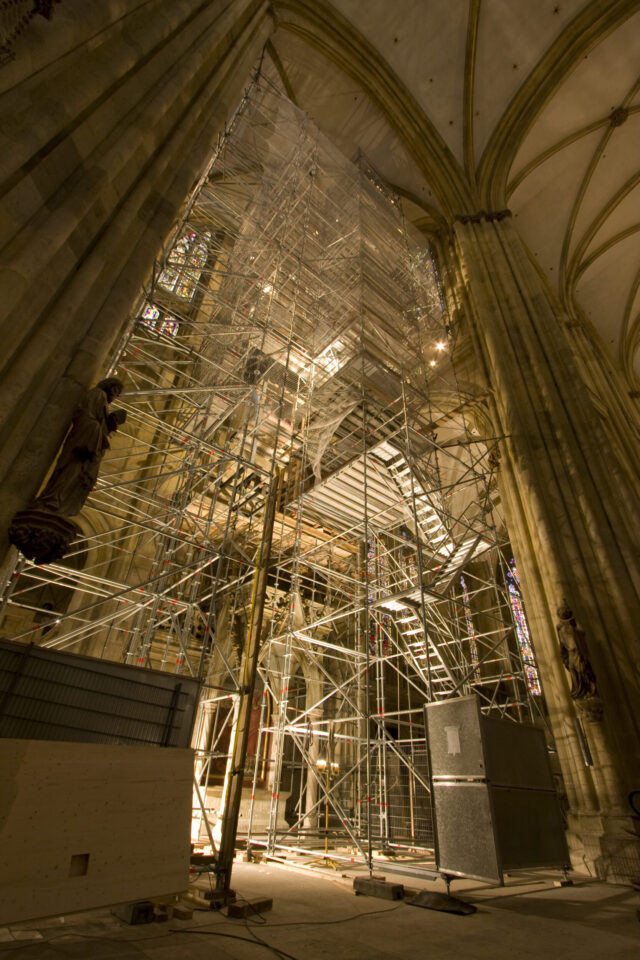 Innenansicht eines großen historischen Gebäudes mit hoher Decke und aufwendiger Architektur; zentrales Gerüst mit mehreren Ebenen und Treppen für Bau- oder Restaurierungsarbeiten, flankiert von Statuen an den Säulen.