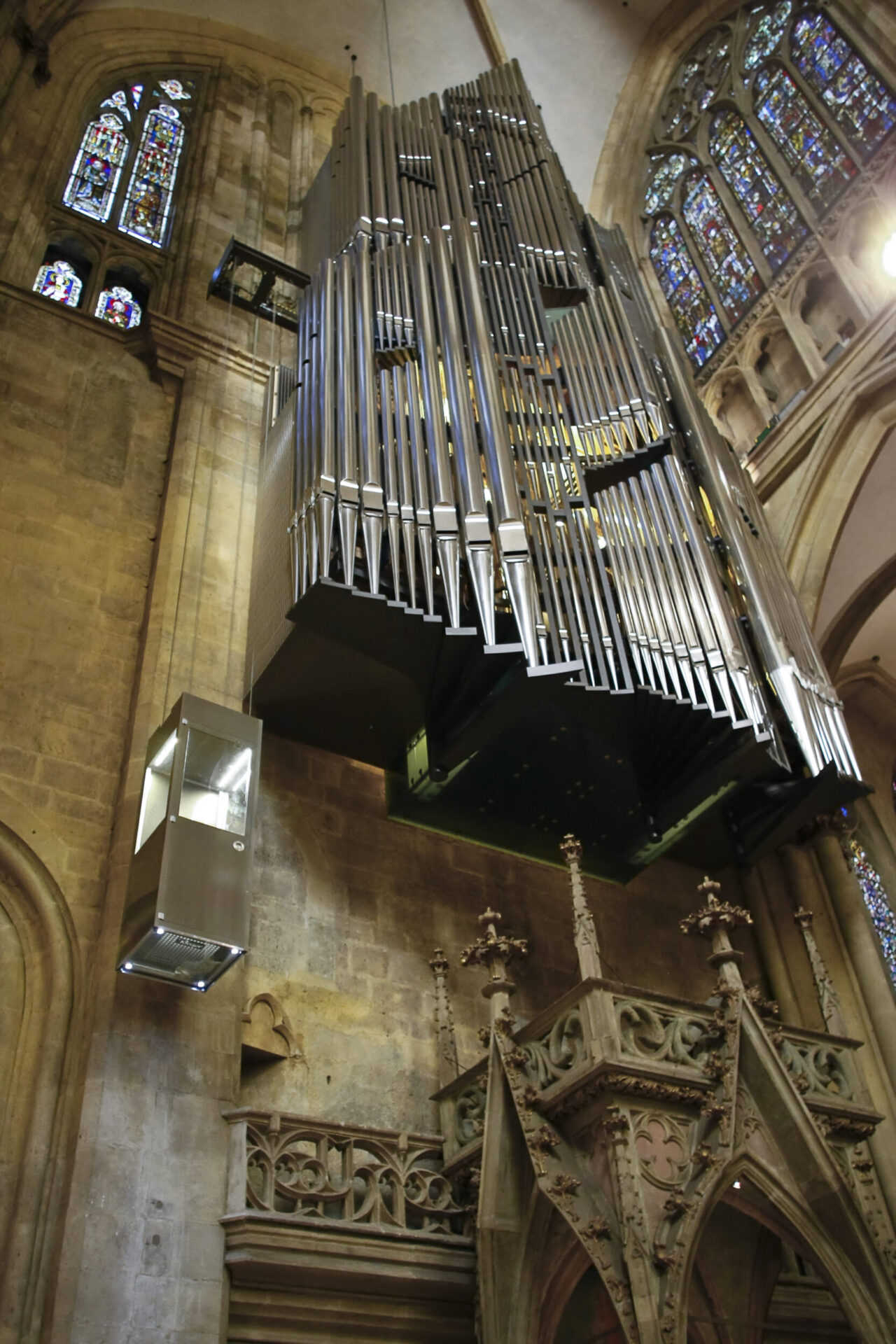 Große Orgel mit zahlreichen Metallpfeifen, hoch an der Wand einer Kirche oder Kathedrale montiert; umgeben von bunten Glasfenstern, steinernen Verzierungen und einem Glas-Metall-Lift an der linken Wand.