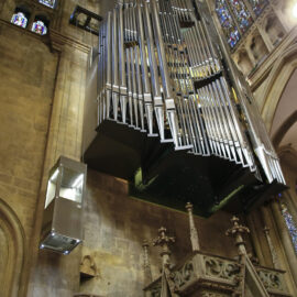Große Orgel mit zahlreichen Metallpfeifen, hoch an der Wand einer Kirche oder Kathedrale montiert; umgeben von bunten Glasfenstern, steinernen Verzierungen und einem Glas-Metall-Lift an der linken Wand.