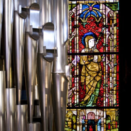 Kircheninnenraum mit großen metallischen Orgelpfeifen auf der linken Seite und einem farbigen Glasfenster mit einer religiösen Figur in Gewändern auf der rechten Seite.