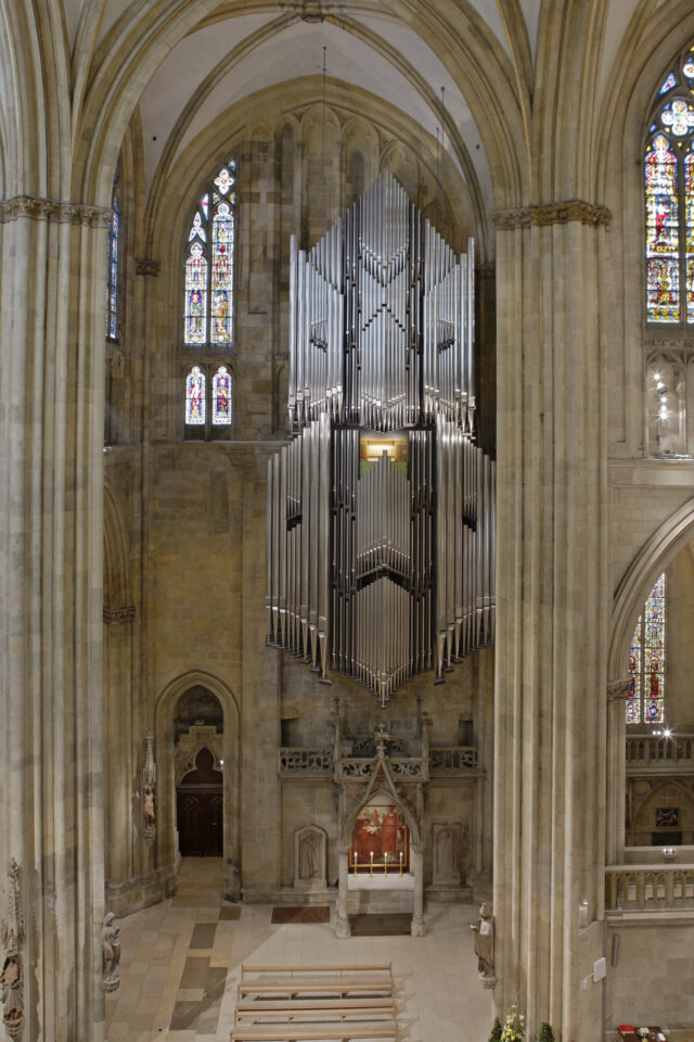 Innenraum einer großen Kathedrale mit hohen Gewölbedecken und bunten Glasfenstern; im Mittelpunkt eine große Orgel mit zahlreichen Metallpfeifen über dem Altar.