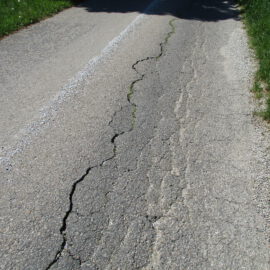 Das Bild zeigt eine Nahaufnahme einer rissigen und abgenutzten Asphaltstraße. Ein großer Riss verläuft längs durch die Fahrbahn, aus dem an einigen Stellen Gras wächst. In der Mitte ist eine weiße gestrichelte Linie zu sehen, und auf beiden Seiten der Straße befindet sich grünes Gras.