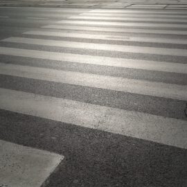 Das Bild zeigt einen Fußgängerüberweg mit weißen Streifen auf einer Asphaltstraße. Sonnenlicht beleuchtet den Übergang und erzeugt Reflexionen auf der Oberfläche. Auf der rechten Seite verläuft ein sichtbarer Riss durch den Asphalt.