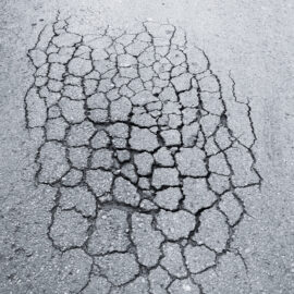 Das Bild zeigt eine stark beschädigte Asphaltfläche mit zahlreichen Rissen. Die Risse verlaufen unregelmäßig und deuten auf eine erhebliche Abnutzung der Straßenoberfläche hin.