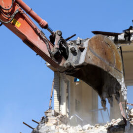 Das Bild zeigt einen Bagger mit einem großen Schaufelaufsatz, der ein Gebäude abreißt. Das Gebäude besteht aus Ziegeln und Beton, und der Bagger reißt aktiv einen Teil der Struktur ein. Rund um die Abrissstelle liegen Trümmer und Bauschutt, im Hintergrund ist ein klarer blauer Himmel zu sehen.