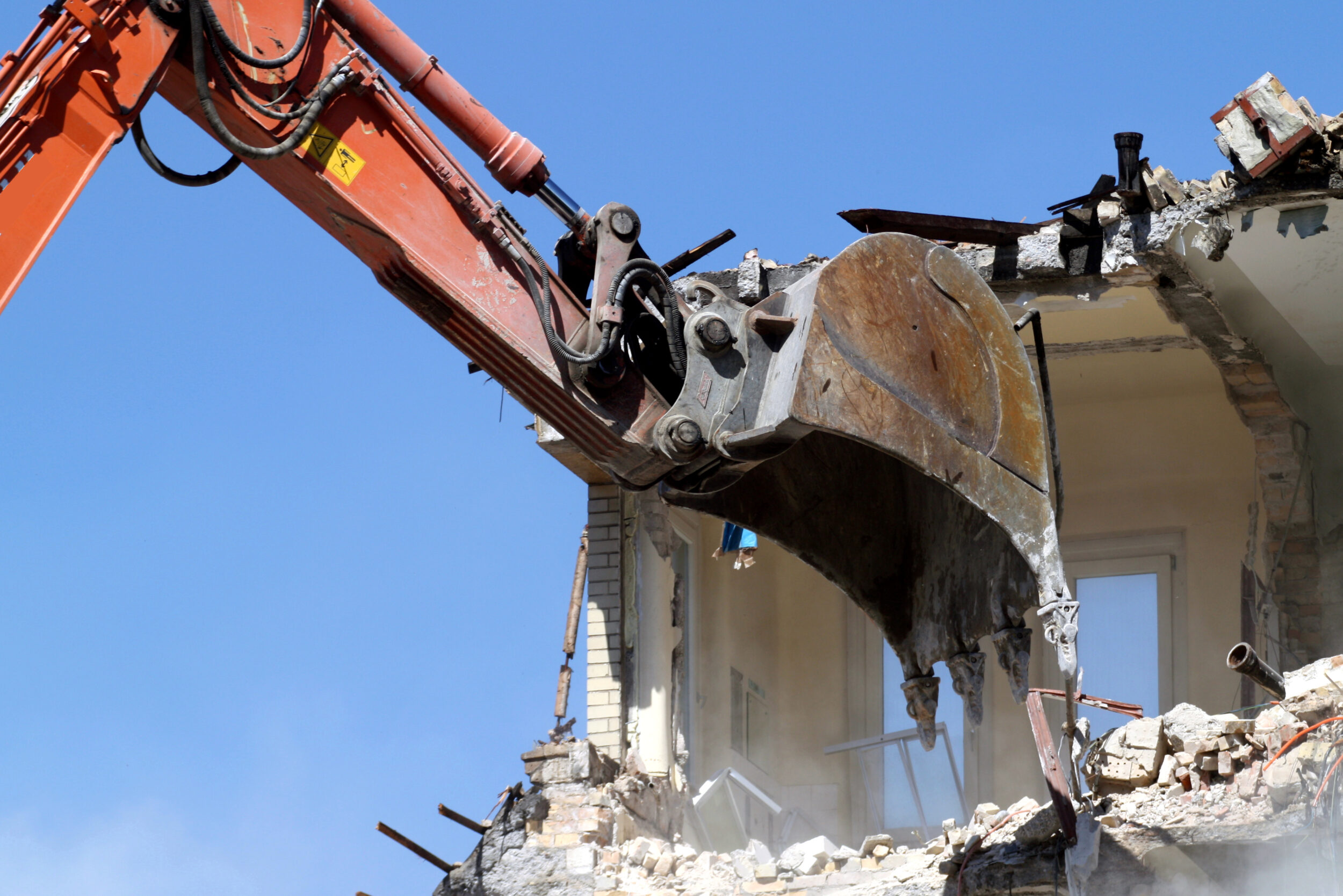 Das Bild zeigt einen Bagger mit einem großen Schaufelaufsatz, der ein Gebäude abreißt. Das Gebäude besteht aus Ziegeln und Beton, und der Bagger reißt aktiv einen Teil der Struktur ein. Rund um die Abrissstelle liegen Trümmer und Bauschutt, im Hintergrund ist ein klarer blauer Himmel zu sehen.