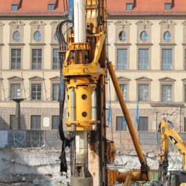 Das Bild zeigt eine Baustelle mit einer großen gelben Bohrmaschine im Vordergrund, die mit hydraulischen Komponenten und Kabeln ausgestattet ist. Im Hintergrund steht ein älteres Gebäude mit mehreren Fenstern und einem roten Ziegeldach. Auf dem Gelände sind außerdem weitere Baumaschinen und Bauschutt zu sehen.