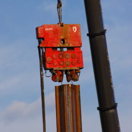 Das Bild zeigt eine Baustelle mit einem schweren roten Vibrationshammer, der an einem Kran befestigt ist. Das Gerät wird verwendet, um einen Stahlträgerpfahl in den Boden zu treiben oder herauszuziehen. Im Hintergrund ist ein klarer Himmel mit einigen Wolken zu sehen.