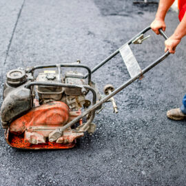 Das Bild zeigt eine Person, die mit einer Rüttelplatte frisch verlegten Asphalt verdichtet. Die Person trägt eine orangefarbene Warnweste und eine blaue Hose. Die Rüttelplatte ist orange und dient dazu, die Asphaltoberfläche gleichmäßig zu verdichten.