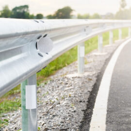 Das Bild zeigt eine Nahaufnahme einer Leitplanke am Straßenrand, die von senkrechten Pfosten getragen wird. An der Leitplanke sind reflektierende Markierungen angebracht, und die Straße verläuft in einer Rechtskurve. Neben der Leitplanke befinden sich Kies und Gras, im Hintergrund sind Bäume und Vegetation unter einem hellen Himmel zu sehen.