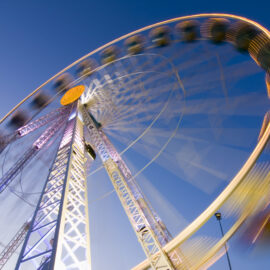 Ein Riesenrad dreht sich vor blauem Himmel. Durch eine Langzeitbelichtung entstehen leuchtende Lichtspuren, die die Bewegung sichtbar machen.
