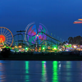 Ein hell erleuchteter Freizeitpark bei Nacht mit Riesenrad, Achterbahn und einem hohen Kettenkarussell. Die bunten Lichter spiegeln sich im Wasser im Vordergrund.