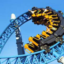 Eine Achterbahn mit gelben und schwarzen Wagen fährt durch eine Looping-Kurve auf einer blauen Schiene. Der Himmel im Hintergrund ist klar und blau.