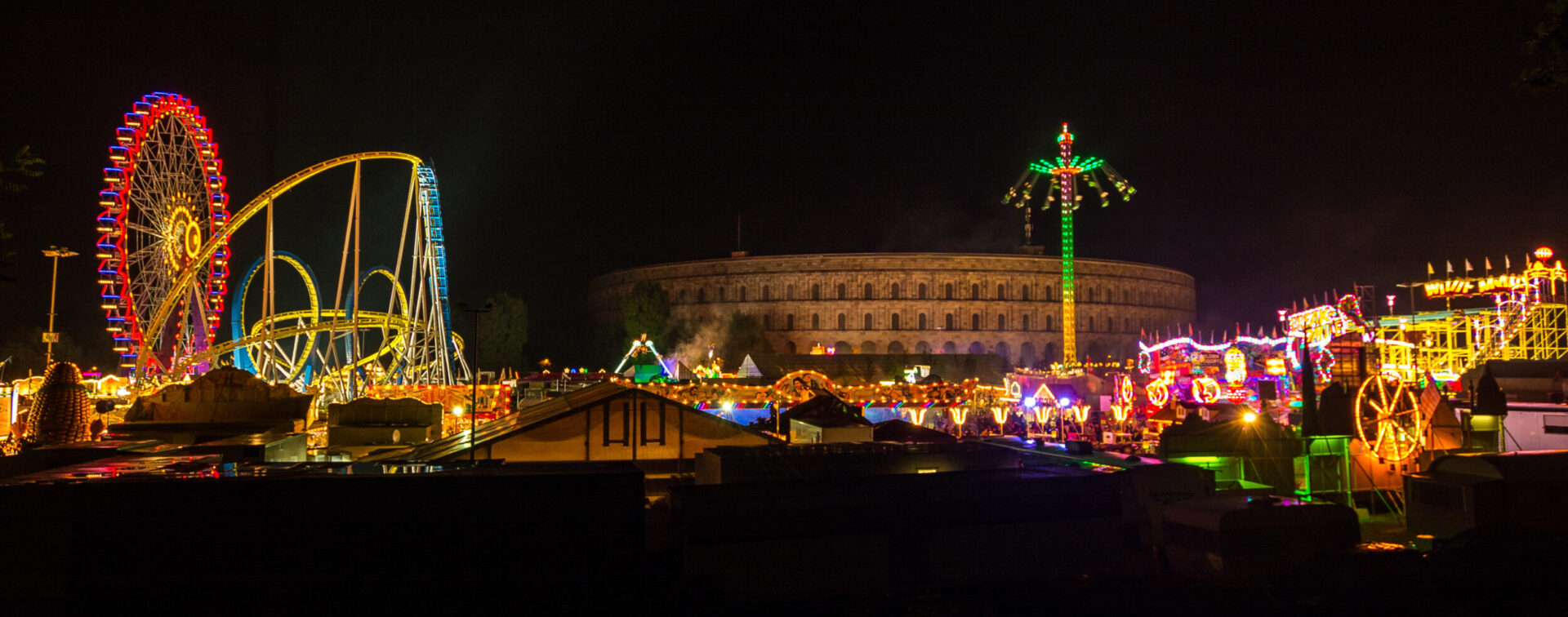 Das Nürnberger Volksfest bei Nacht mit beleuchtetem Riesenrad, Achterbahn mit gelben Schienen und weiteren Fahrgeschäften. Im Hintergrund ist das Kolosseum zu erkennen.