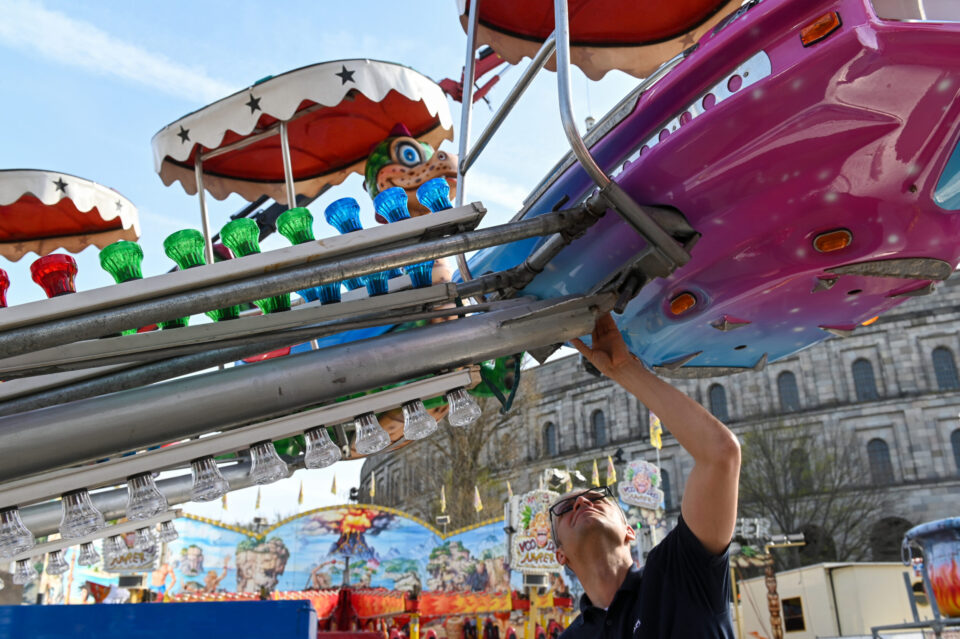 Person bei der Inspektion oder Wartung eines Fahrgeschäfts mit bunten Lichtern und einem pink-lila Sitz, im Hintergrund weitere Attraktionen und Strukturen des Nürnberger Volksfest.