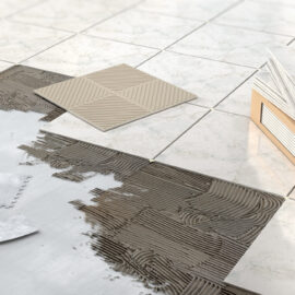 Das Bild zeigt eine Szene beim Verlegen von Fliesen. Mehrere Fliesen sind bereits auf dem Boden platziert, und auf einer freien Fläche ist Kleber aufgetragen, bereit für weitere Fliesen. Ein Zahnspachtel und ein Spachtel liegen auf dem Kleber, daneben steht ein Karton mit zusätzlichen Fliesen, von denen eine angelehnt und eine flach auf dem Boden liegt. Die Fliesen haben eine marmorartige Optik mit hellgrauer Maserung.