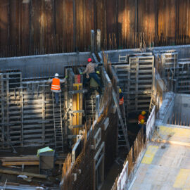 Das Bild zeigt eine Baustelle mit mehreren Arbeitern, die Schutzkleidung wie Helme und Warnwesten tragen. Die Arbeiter sind mit dem Bau einer Betonstruktur beschäftigt, umgeben von Metallgerüsten und Holzbohlen. Auf dem Gelände liegen verschiedene Baumaterialien, und die Baustelle ist von hohen Metallwänden eingefasst.