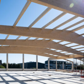 Das Bild zeigt den Rohbau einer Dachkonstruktion mit gebogenen Holzbalken, die von senkrechten Betonsäulen getragen werden. Auf der rechten Seite sind Gerüste und Baumaschinen zu sehen. Im Hintergrund befinden sich Gebäude und Bäume unter einem klaren blauen Himmel.