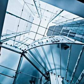 Das Bild zeigt eine moderne Glasarchitektur mit einer spiralförmigen Treppe, die nach oben führt. Umgeben von Glaswänden und einer gläsernen Decke entsteht ein transparenter, lichtdurchfluteter Raum. Im Hintergrund ist durch das Glas ein hohes Gebäude zu erkennen.