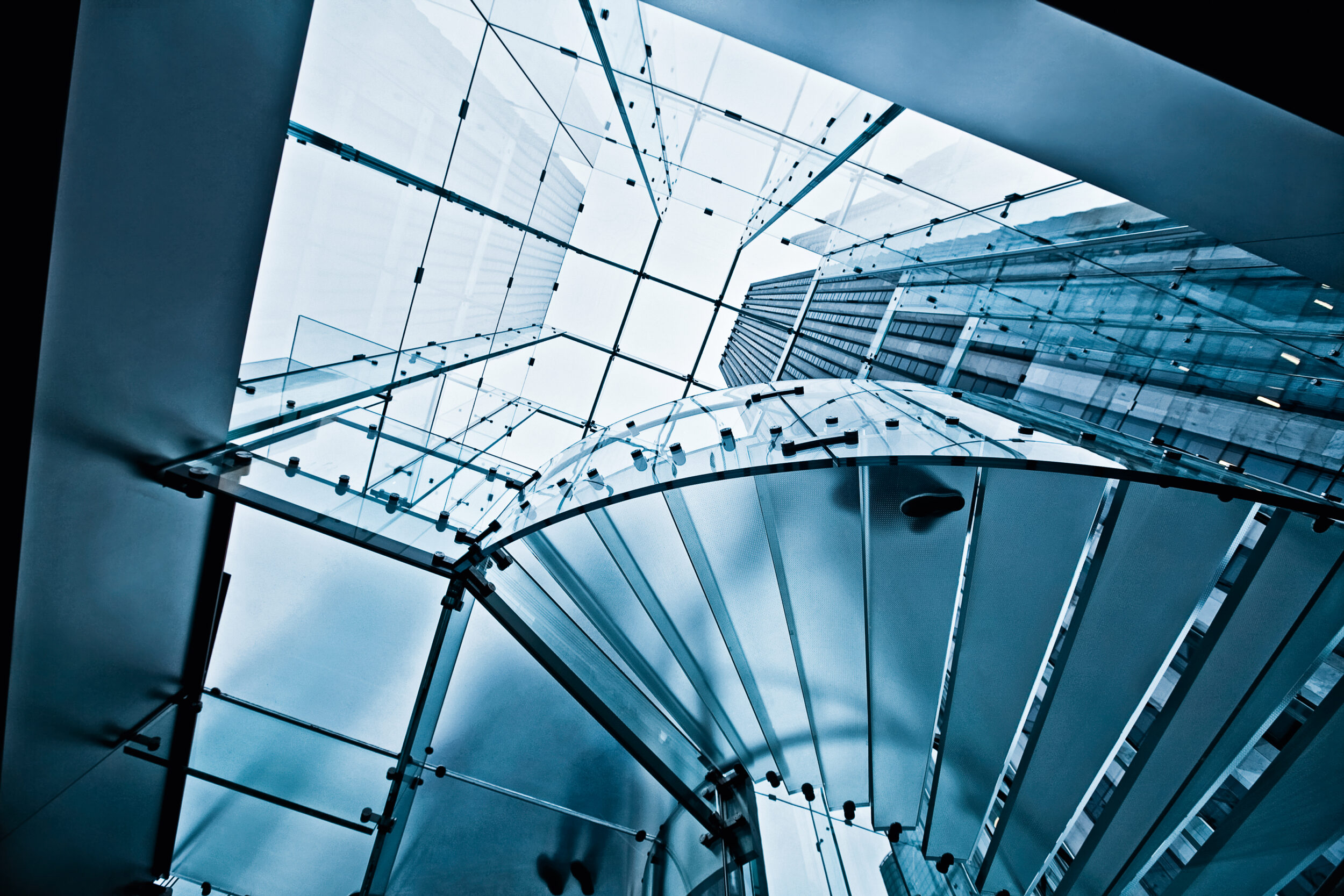 Das Bild zeigt eine moderne Glasarchitektur mit einer spiralförmigen Treppe, die nach oben führt. Umgeben von Glaswänden und einer gläsernen Decke entsteht ein transparenter, lichtdurchfluteter Raum. Im Hintergrund ist durch das Glas ein hohes Gebäude zu erkennen.