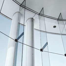 Das Bild zeigt eine moderne Architektur mit großen Glasflächen, die von Metallhalterungen und Seilen getragen werden. Die Glasstruktur ist gebogen und wird von weißen Säulen sowie einer Decke mit eingelassenen Leuchten ergänzt. Durch die Glasflächen ist der Himmel sichtbar, was auf einen Außenbereich oder ein Atrium hinweist.