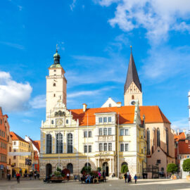 Belebter Stadtplatz in Ingolstadt mit dem historischen Alten Rathaus und Uhrturm im Zentrum, umgeben von Gebäuden mit roten Ziegeldächern und moderner Architektur, unter blauem Himmel mit vereinzelten Wolken.