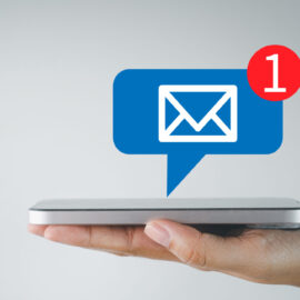Hand mit Smartphone, über dem ein Benachrichtigungssymbol in Form einer blauen Sprechblase mit Briefumschlag und rotem Kreis mit der Zahl „1“ schwebt, was auf eine neue Nachricht hinweist.