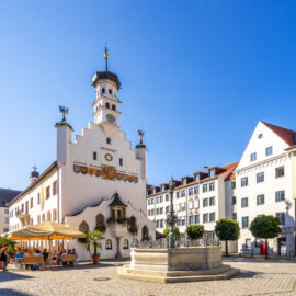 Belebter Stadtplatz in Kempten mit einem historischen Gebäude samt Uhrturm und verzierten Fassadendetails, davor Außengastronomie mit Sonnenschirmen sowie ein steinerner Brunnen rechts, umgeben von Bäumen und traditionellen Gebäuden unter blauem Himmel.