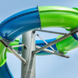 Das Bild zeigt eine Nahaufnahme einer Wasserrutsche mit zwei ineinander verschlungenen Röhren, eine blau und eine grün. Die Röhren sind glänzend und werden von Metallstreben an einem zentralen Mast gehalten. Im Hintergrund ist ein klarer, blauer Himmel zu sehen.