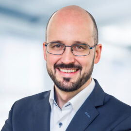 Andreas Kupzok trägt eine Brille, ein Sakko und ein Hemd mit Kragen. Der Hintergrund ist unscharf und zeigt ein Büro.