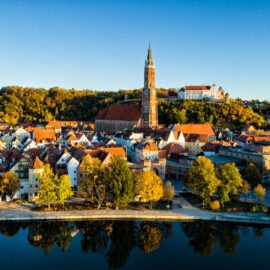 Luftaufnahme von Landshut in Bayern mit der markanten St. Martinskirche im Zentrum, umgeben von roten Ziegeldächern und bunten Gebäuden, im Hintergrund bewaldete Hügel mit Herbstlaub und ein Fluss im Vordergrund.