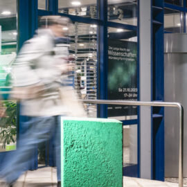 Eine Person betritt durch einen Eingang mit grünem Licht eine Veranstaltung; im Hintergrund ist ein Plakat zur „Langen Nacht der Wissenschaften“ am 21.10.2023 in Nürnberg, Fürth und Erlangen zu sehen.