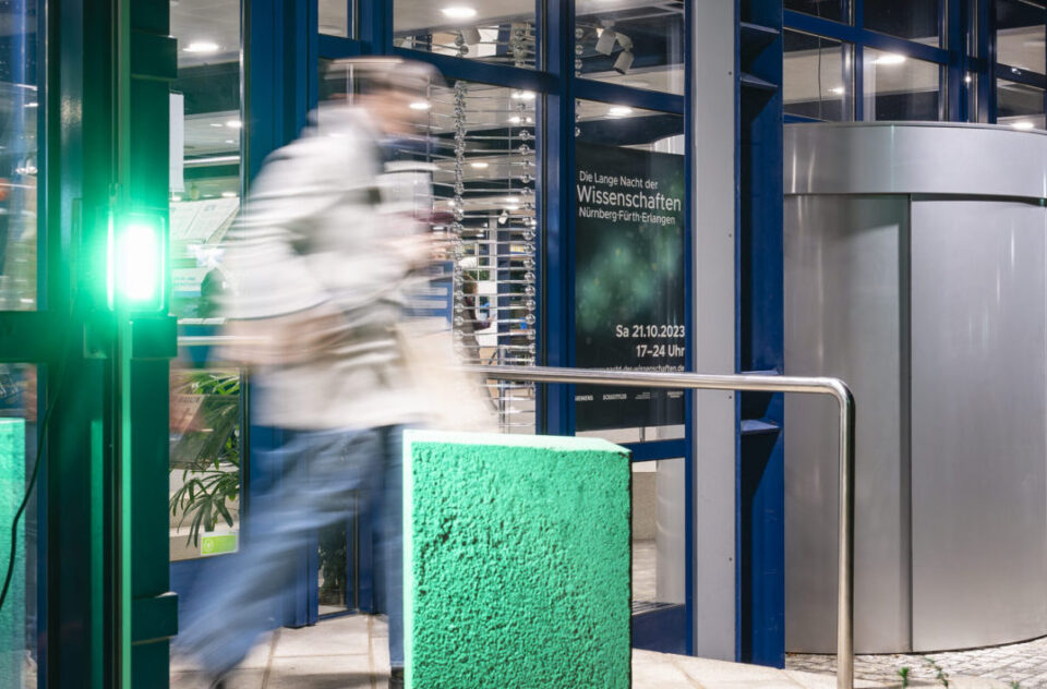 Eine Person betritt durch einen Eingang mit grünem Licht eine Veranstaltung; im Hintergrund ist ein Plakat zur „Langen Nacht der Wissenschaften“ am 21.10.2023 in Nürnberg, Fürth und Erlangen zu sehen.