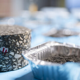 Ein zylindrisches Asphaltprobenstück mit sichtbaren Gesteinskörnungen und roter Markierung liegt neben einer Aluminiumschale mit dunklem Material; im Hintergrund sind weitere Proben auf einer blauen Fläche zu erkennen.