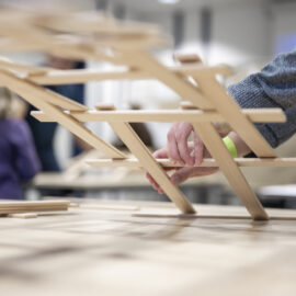 Nahaufnahme von Händen, die aus Holzstäbchen eine brückenartige Konstruktion bauen; im unscharfen Hintergrund sind weitere Personen und Gegenstände zu erkennen.