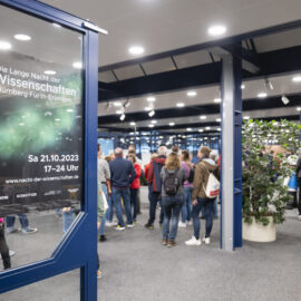 Mehrere Personen befinden sich in einem Gebäude bei einer Veranstaltung; auf einer Tür ist ein Plakat zur „Langen Nacht der Wissenschaften“ am 21.10.2023 mit Veranstaltungsorten und Uhrzeit zu sehen.