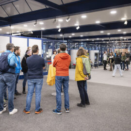 Menschen befinden sich in einem hell erleuchteten Foyer bei einer Veranstaltung; einige stehen an einem Tisch mit gelber Tischdecke, andere bewegen sich im Raum, rechts ist etwas Grünpflanzen-Dekor zu sehen.