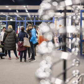 Menschen befinden sich in einem öffentlichen Innenraum mit Glaswänden und Plakaten; im Vordergrund ist unscharf ein Modell zu sehen, das einer DNA-Doppelhelix ähnelt.