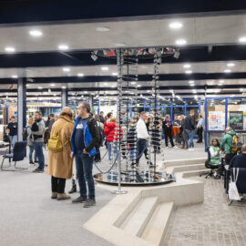 Menschen befinden sich bei einer Veranstaltung in einem öffentlichen Innenraum mit Glaswänden und Plakaten; im Vordergrund ist ein metallisches Modell zu sehen, das einer DNA-Doppelhelix ähnelt.