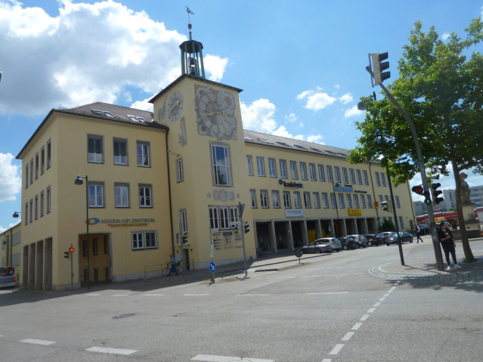 Ansicht des gelben Gebäudes mit Uhrturm in Ansbach, mit parkenden Autos und einer Straßenkreuzung im Vordergrund bei leicht bewölktem Himmel.