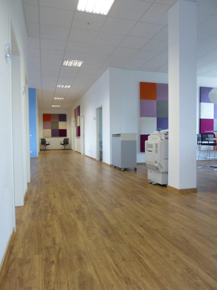 Moderner Büroflur mit Holzfußboden, weißen Wänden und farbigen Wandpaneelen, einem Drucker auf der rechten Seite sowie einem offenen Bereich mit hohen Stühlen und Tischen im Hintergrund.